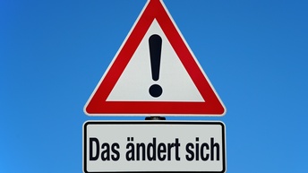 Zu sehen ist ein blauer Hintergrund mit einem Warnschild. Unter dem Schild ist ein Schild mit der Benennung 