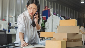 Person telefoniert und notiert sich gleichzeitig etwas in einem Notizbuch, während Kartonagen sowie ein Tablet in einem Lagerraum sowie einer Produktionsstätte mit Nähmaschinen stehen