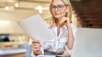 Eine Person mit Brille und in einer Bluse sitzt an einem Tisch in einem Wohnessbereich. Die Person hält einen Zettel, auf den sie blickt. Vor ihr steht ein aufgeklappter Laptop. Im Hintergrund sind eine Küche und eine Treppe