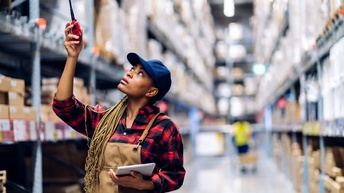 Person mit blauer Kappe und Arbeitslatzhose in Gang zwischen Regalen in Lagerhalle stehend hält einen Arm nach oben mit WalkieTalkie in der Hand, in anderem Arm Tablet haltend, im Hintergrund verschwommen weitere Person und Lagerregale