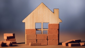 Auf einer hellen Oberfläche steht eine helle Häuserfront in einer Modellform. Teilweise sind vor der Häuserfront kleine Ziegelsteine. Um die Häuserfront liegen gestapelte und lose Ziegelsteine.