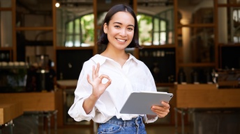 Lächelnde Person mit dunklen Haaren weißer Bluse und  Jeans steht mit einem Tablet vor einem Lokal und gibt ein Handzeichen für Zustimmung