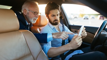 Zwei Personen in Fahrzeugsitzend blicken auf Smartphone, eine Person deutet mit Finger auf Device