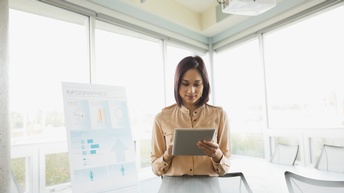 Frontale Aufnahme einer Person in Bürokleidung, die in einem Raum mit Glasfronten steht und auf ein Tablet blickt, das sie hält. Links hinter der Person steht ein Plakat, auf dem Infografiken steht. Rechts steht ein großer Tisch mit Sesseln