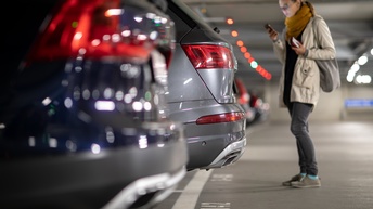 Parkende Autos mit Fokus auf Heck in einer Parkgarage und eine Person, die dahinter steht und auf ein Smartphone blickt