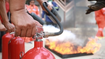 In der linken Bildhälfte sind mehrere Feuerlöscher. Auf den Feuerlöschern sind die Hände von Personen. Die Personen halten die Schläuche der Feuerlöscher auf ein Feuer, das im Hintergrund ist