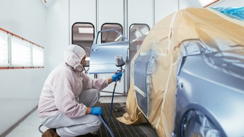Person in weißer Arbeits- und Schutzbekleidung mit Atemmaske und blauen Handschuhen lackiert ein Auto mit einem Sprüh-Kompressor