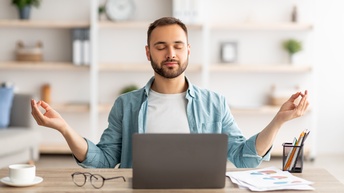 Person mit T-Shirt und blauem offenen Hemd sitzt an einem Schreibtisch mit meditativer Pose während am Schreibtisch ein Laptop, eine Brille, eine Tasse, ein Stiftehalter sowie Unterlagen liegen und im Hintergrund ein Regal in der Unschärfe steht