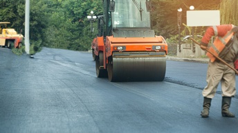 Auf einer frisch asphaltierten Straße steht ein Fahrzeug mit einer Walze vorne. Daneben steht eine Person in Stiefeln und oranger Jacke