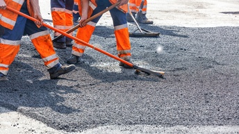 Mehrere Personen in oranger Arbeitskleidung und dicken Arbeitsschuhen bearbeiten mit Rechen und Schaufeln eine asphaltierte Straße