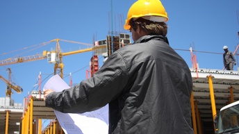 Rückenansicht einer Person mit Helm, die einen Bauplan hält. Im Hintergrund ist eine Baustelle eines Gebäudes sowie stehen zwei Baukräne