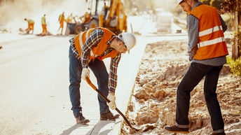 Zwei Personen mit Sicherheitsweste, Helm und Handschuhen schaufeln mit Schaufeln Erde vom Straßenrand weg. Im Hintergrund stehen weitere Personen und ein Baumaschinengerät