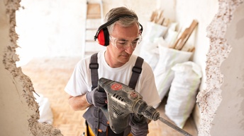 In einer Wand in einem Wohnraum ist ein großes, ausgefranstes Loch. Eine Person mit Handschuhen, Schutzbrille und Gehörschutz erweitert das Loch mit einer Bohrmaschine. Hinter der Person stehen Säcke mit Schutt sowie eine Leiter