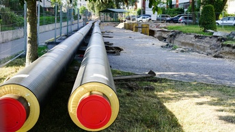 Zwei lange Rohre liegen teilweise auf grüner Wiese, teilweise auf einer Straße. Links von ihnen ist ein langer Metallzaun. rechts von ihnen ist ein kleiner Graben