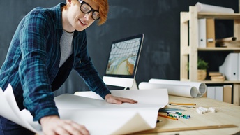 Lächelnde Person mit Mütze und Brille beugt sich über Tisch und breitet weißen Papierbogen aus, im Hintergrund verschwommen Computermonitor und Regal mit Ordnern