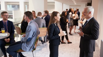 Personen in Businesskleidung stehen bei einem Empfang zusammen und unterhalten sich, einige halten Kaffeetassen, Saftgläser oder ein Teller mit Speisen in Händen
