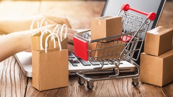 Auf einem Tisch steht ein Miniatur-Einkaufswagen, in dem zwei kleine Kartons stehen. Neben dem Wagen stehen zwei weitere Kartons und kleine Einkaufstaschen. Dahinter sind die Hände einer Person auf der Tastatur eines Laptops.