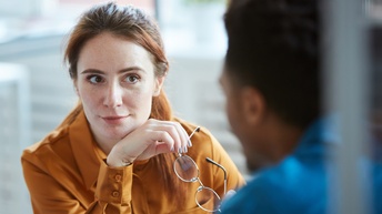 Person im Fokus mit rotbraunen langen Haaren und senfgelber Bluse hält Brille in der Hand und blickt interessiert zu einer anderen Person, die in Unschärfe dargestellt ist