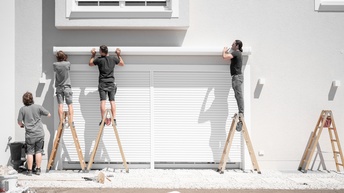 Drei Personen auf Holzstehleitern halten Markisenpaneelzur Befestigung über bodentiefe Fenster mit heruntergelassenen Außenjalousien an weiße Hausfassade, weitere Person am Boden stehend blickt zu ihnen hinauf