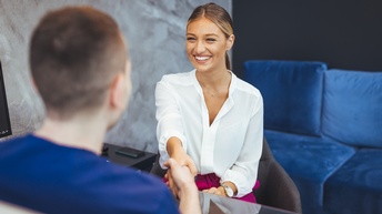 Rückenansicht einer Person, die einer anderen Person an einem Tisch gegenüber sitzt. Die Personen schütteln sich die Hände. Die zur Kamera gewandte Person lächelt