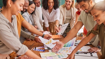 Mehrere Personen stehen freudig rund um einen Besprechungstisch, halten und zeigen auf Farbkarten und weitere Unterlagen