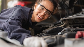 Nahaufnahme einer Person mit Schutzbrille und Handschuhen, die sich mit dem Oberkörper nach vorne über einen Motor beugt und direkt in die Kamera blickt