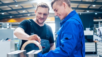 xt<br />
Zwei Personen in Overalls im Werkshalle stehend im Gespräch miteinander: Eine Person hält breites metallenes Verbundstück in Händen und blickt darauf sprechend