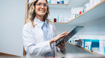 Eine Person mit weißem Kittel, Brille und schulterlangen, hellen Haaren steht in einem Raum mit Regalen an den Wänden und hält ein Tablet mit der linken Hand. Auf den einzelnen Regalböden liegen Medikamentenschachteln
