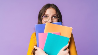 Frontale Aufnahme einer Person mit langen, dunklen Haaren und Brille vor einem lila Hintergrund hält in den Händen vier bunte Hefte und blickt nach oben. Ihre Hände sind verschränkt und im Fokus des Bildes