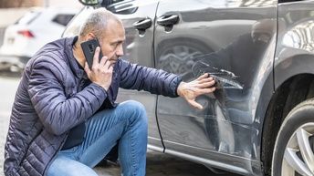 Eine Person hockt neben einem dunklen, geparkten Auto, das eine Delle und einen Kratzer hat. Die Person telefoniert und blickt auf den Schaden