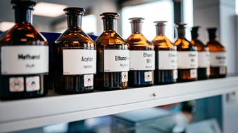 Braune Glasflaschen mit weißen Etiketten und schwarzen Aufschriften nebeneinander in Regal stehend, im Fokus Apothekerflasche mit Schriftzug Ethanol und Symbol einer Flamme