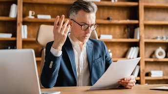 Person im Anzug trägt eine Brille und sitzt mit fragendem Blick an einem Schreibtisch mit Computer und hält Papier in einer Hand ein Papier und in der anderen einen Stift, im Hintergrund steht ein braunes Holzregal mit Büchern und Dekoelementen