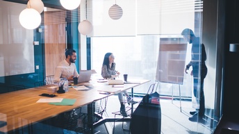 Zwei Personen befinden sich in einem gläsernen Besprechungsraum bei einem Besprechungstisch mit Unterlagen und Laptop, eine Person steht bei einem Flipchart