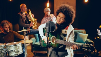 Im Fokus Person vor Mikrofon sitzend singt und spielt Gitarre, im Hintergrund weitere Personen unterschiedliche Musikinstrumente spielend wie Schlagzeug und Saxophon