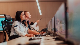 Person in Arbeitskleidung sitzt an einem Schreibtisch und telefoniert während daneben weitere Personen sitzen
