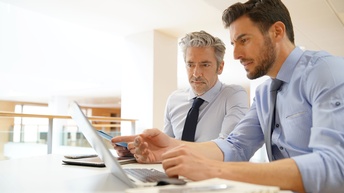 Zwei Personen in Hemden und mit Krawatten sitzen nebeneinander an einem Tisch. Vor ihnen steht ein aufgeklappter Laptop, auf den beide klicken. Neben dem Laptop sind ein Notizbuch und ein Tablet