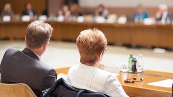 Rückenansicht von zwei Personen in Sakkos, die nebeneinander an einem Tisch sitzen. Im Hintergrund ist ein sehr langer Tisch, an dem viele verschiedene Personen sitzen