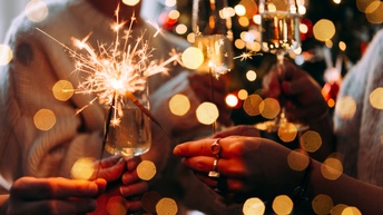 Nahaufnahme der Hände von drei Personen, die in einem Kreis stehen. Jede Person hält in einer Hand ein Sektglas mit Sekt und in der anderen Hand eine Wunderkerze