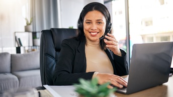 Frontalaufnahme einer an einem Tisch sitzenden Person. Die Person trägt ein Headset. Vor ihr ist ein aufgeklappter Laptop. Die Person blickt direkt in die Kamera
