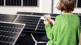 Person in grünem Pullover in Rückenansicht steht inmitten einer Solaranlage und hält Fächer mit Eurogeldscheinen in Händen, im Vordergrund Modell eines Windrades
