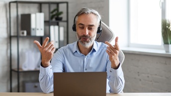 Person mit kurzen grauen Haaren, Bart und hellblauem Hemd sitzt an einem Schreibtisch in einem Büro, trägt kabellose Kopfhörer und blickt sowie gestikuliert mit den Händen in einen aufgeklappten Laptop