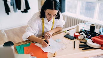 Person mit kurzen dunklen Haaren und Brille sowie weißer Bluse mit umgehängten Maßband sitzt an einem Tisch und zeichnet ein Schnittmuster, am Tisch liegen weitere Nähutensilien, im Hintergrund steht ein voller Kleiderständer