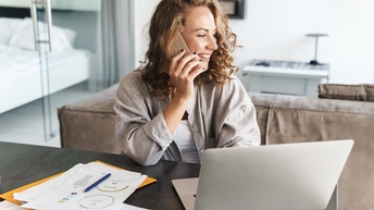 Eine Person mit lockigem Haar sitzt an einem Tisch in einem Wohnessbereich und telefoniert mit dem Handy. Vor ihr auf dem Tisch sind ein aufgeklappter Laptop und verschiedene Zettel mit Diagrammen und Zahlen