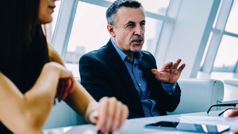 Zwei Personen sitzen nebeneinander an einem Tisch. Vor ihnen sind ein Tablet und eine Brille. Hinter ihnen ist eine leicht schräge Fensterfront. Die rechte Person gestikuliert mit der rechten Hand und spricht.