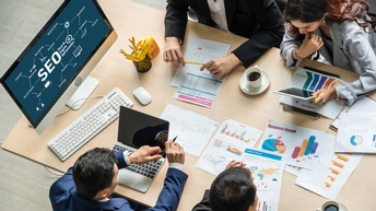 Personen in Businesskleidung sitzen bei einem Besprechungstisch und Unterlagen sowie Laptops liegen darauf, am Computerbildschirm wird ein SEO Konzept visualisiert, Vogelperspektive