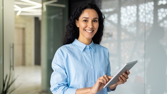 Person mit dunklen langen Haaren und blauer Bluse blickt freudig in die Kamera während ein Tablet mit beiden Händen gehalten wird, im Hintergrund zeigt sich ein offener Flur
