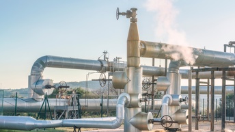 In einem eingezäunten Bereich ist ein Teil asphaltiert. Daneben ist grünes Gras. Auf dem asphaltierten Bereich verlaufen Pipelines in unterschiedlichen Höhen und verschiedenen Breiten. Der Himmel ist blau. An einer Stelle steigt Dampf aus