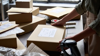 Auf einem Tisch liegen mehrere Pakete, Kuverts sowie durchsichtige Folie. Eine Person steht seitlich vor dem Tisch und hat ihre beiden Händen auf einem Paket