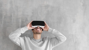 Person in hellgrauem Pullover steht vor einer <br />
Betonwand und trägt eine Virtual Reality Brille 