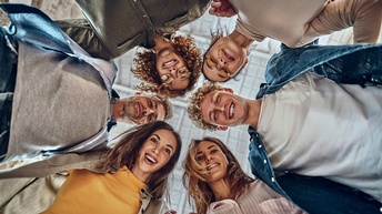 Aufnahme aus der Froschperspektive von sechs in einem Kreis stehenden Personen, die nach unten zur Kamera blicken. Im Hintergrund ist die Decke eines Raums